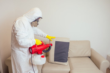 pest control worker in uniform spraying pesticides under couch in living lounge room