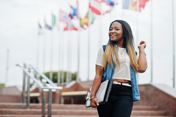 African student female posed with backpack and school items on yard of university, against flags of...