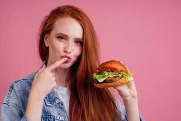 beautiful redhaired ginger redhead woman holding big huge cheeseburger with cheese,beef cutlet and tomato lettuce and licking fingers in studio pink background