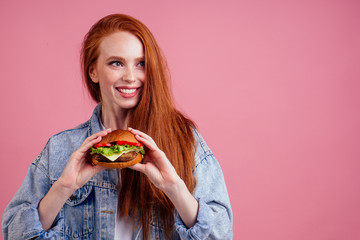 beautiful redhaired ginger redhead woman holding big cheeseburger with cheese,beef cutlet and tomato lettuce in studio pink background