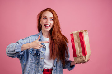 happy long red-haired ginger woman holding paper eco bag package with chips in studio pink background