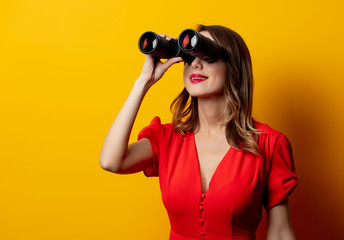 Young woman in red dress with binocular on yellow background