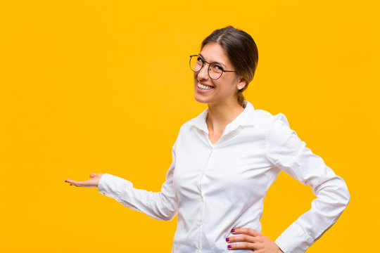 young businesswoman feeling happy and cheerful, smiling and welcoming you, inviting you in with a friendly gesture against orange wall