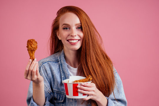 pretty long redhead ginger girl eating fries chicken in studio pink background