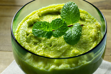 closeup of avocado dessert in green glass jar with mint on top over wooden table
