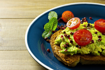 avocado cream toast with sliced tomatoes and pepper on top in blue plate garnished with mint on wooden table