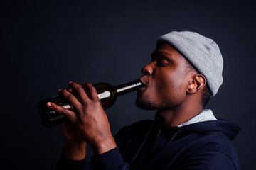 african american gue wearing gray hat and hoody hjlding botle with wine in srudio black background.alcoholism homeless bum concept