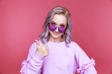 Portrait of young woman in bright glasses showing thumb sign standing isolated over the pink background