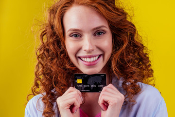 attractive redhead curly ginger caucasian businesswoman wearing cotton shirt ,holding credit card studio yellow background ,money for for dental treatment braces and whitening