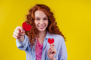 Happy redhead ginger curly girl businesswoman in a cotton shirt model hold decorative hearts Valentines Day gift on yellow background studio.sex education