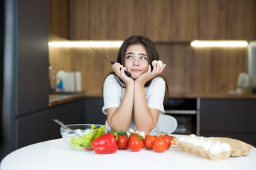 young beautiful woman holding knife in one hand and mushroom in another cooking breakfast in the kitchen in a looking dreamy