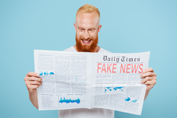 cheerful bearded man reading newspaper with fake news, isolated on blue