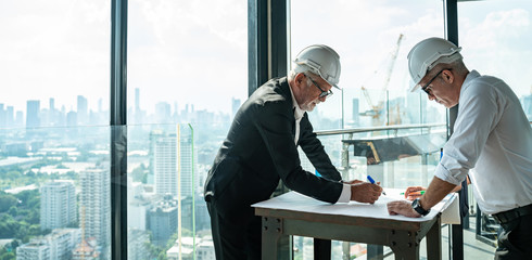 Two Engineer or Architect are analyzing blueprints while working on a new project on construction site with blue sky and city background.Architect supervising construction on terrace tower.