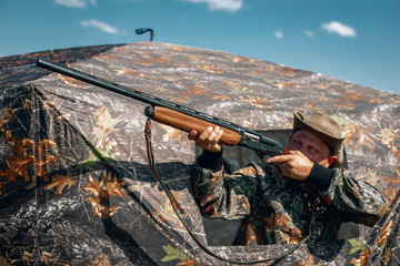 Man standing in ambush in the khaki tent, aiming to the sky; huntsman concept.