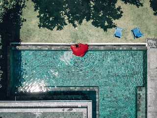 Aerial view of the stunning girl in the pool be the hotel. The young lady wears red dress that flows in the water; fashion concept.