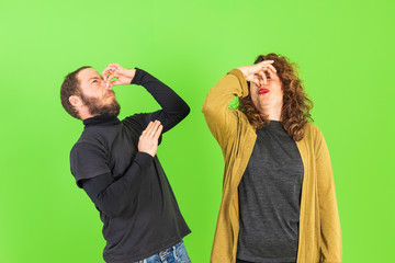 Young beautiful couple on isolated green background smelling something stinky and disgusting, intolerable smell, holding breath with fingers on nose. Bad smells concept.