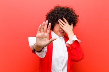 young pretty afro woman covering face with hand and putting other hand up front to stop camera,...