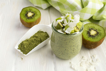 Pudding with chia, matcha tea, kiwi and coconut flakes in the glass jar on the white wooden...