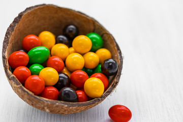 Round balls of milk chocolate covered with multi-colored glaze lie in half a coconut shell on a white background, side view from above