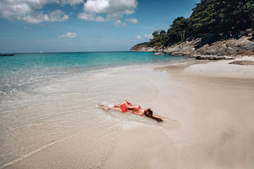 Fototapeta na wymiar Sexy girl in red bikini sunbathing on wet sand by the clear ocean, sunny day, rocks, trees; sunbathing concept.
