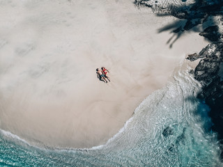 Aerial view of the loving couple lying on the sandy beach, turquoise water; honeymoon concept.