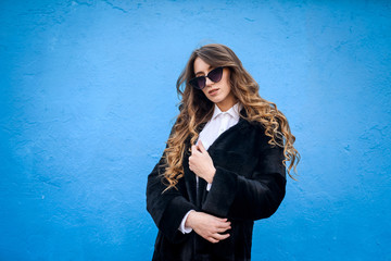 Stylish young woman in fur coat on blue wall background. Beauty and fashion.