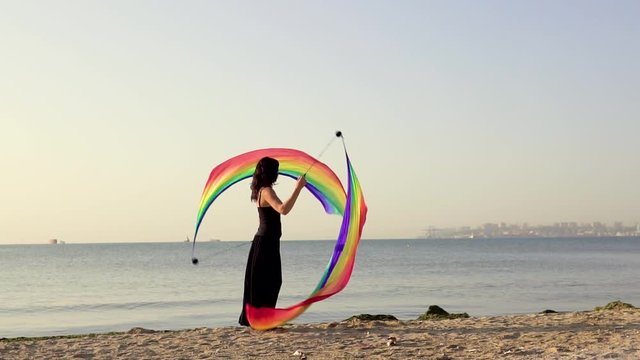Beautiful young girl juggles poi with the LGBT flag on the beach near sea water at sunset. Sleight of hand and acrobatic with poi. slow motion.