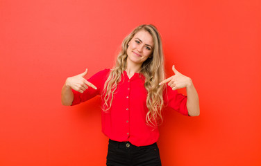 young blonde woman looking proud, arrogant, happy, surprised and satisfied, pointing to self,...
