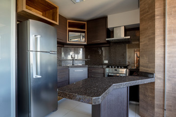 Kitchen with silver fridge, stove and microwave. Central countertop. Dark environment with black granite furniture and dark wood.