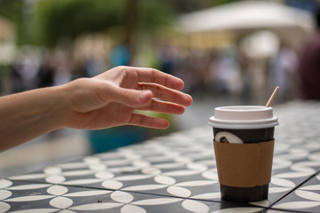 A hand reaches for dark vending coffee in disposable kraft paper cup and plastic lid on table at street cafe.