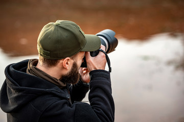 Man photographer holding camera with long lens and taking pictures in outdoors during sunny autumn day.