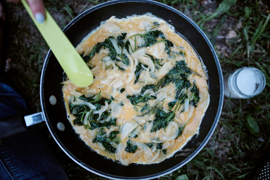 Top view of a woman cooking an omelette with spinach and onions using a camping fire 