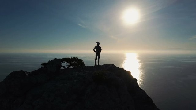 Aerial silhouette of young Successful confident woman mountaineer on mountaintop in Crimea with arms outstretched. Brave Woman standing on the top of a mountain against the sea at sunset.