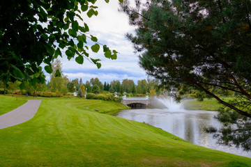 Fototapeta na wymiar Landscape. Hills with green lawn and ornamental shrubs and trees lake fountain and bridge. Beautiful garden