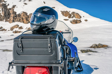 Snowmobile safety helmet lays at the luggage trunk mounted on backside of blue snowmobile, close up view. Blurry mountains covered by snow at the background. Forbidden to drive without helmet, Sweden
