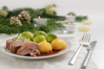 Seared duck breast fillet with duchess potatoes and brussels sprout,  a festive dinner served with candles, fir branches and Christmas decoration on a white wooden table
