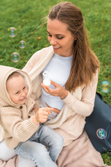 Happy family mom woman little boy weekend resting, laughing, smiling, having fun, playing. Blow soap bubbles, background green grass. Warm casual wear, sweater with hood. Caring support love nurture.