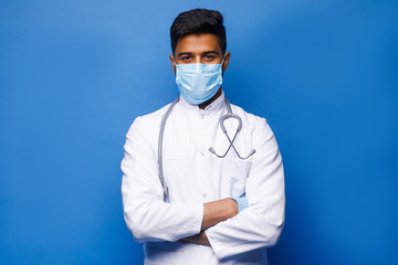 A indian surgeon standing setting his gloves to start the surgery, wearing a mask and a blue uniform, isolated on a blue background.