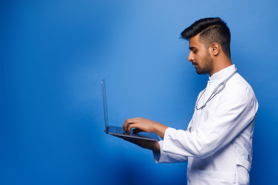 Profile Photo, Indian Male Doctor Using Laptop, Standing Isolated Over Light Blue Colour Background.