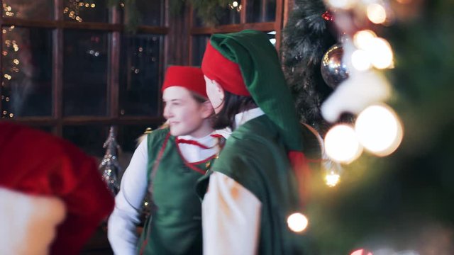 2 Christmas elves are dancing near the window in a room with New Year's decorations.