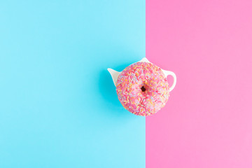 Creative pattern of donut covered with pink icing and colored pastry topping on a plate in the form of a teapot on a pink and blue pastel colours background. Copy space.