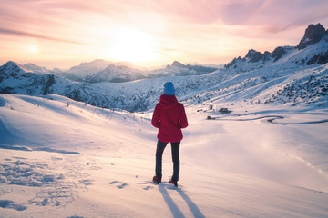 Young woman in snowy mountains at sunset in winter. Landscape with beautiful slim girl on the hill against snow covered rocks and colorful sky with clouds in the evening. Travel in Dolomites. Tourism.