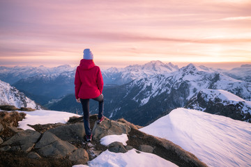 Young woman in snowy mountains at sunset in winter. Beautiful slim girl on the mountain peak against snow covered rocks and colorful red sky with clouds in the evening. Travel in Dolomites. Tourism