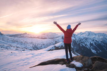 Happy young woman in snowy mountains at sunset in winter. Beautiful slim girl on the mountain peak with raised up arms, snow covered rocks and colorful sky with clouds. Travel in Dolomites. Tourism - Powered by Adobe