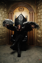 Dark Angel man with huge wings in black outfit sitting on the throne on golden gates background...