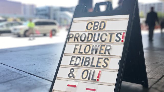A daytime view of a sign outside of a CBD products store. Traffic and pedestrians pass by in the background.  	