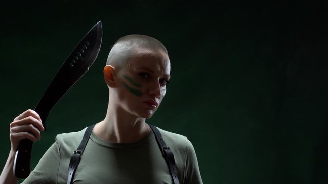 Angry bald girl with camouflage makeup shaving machete hair on a black background