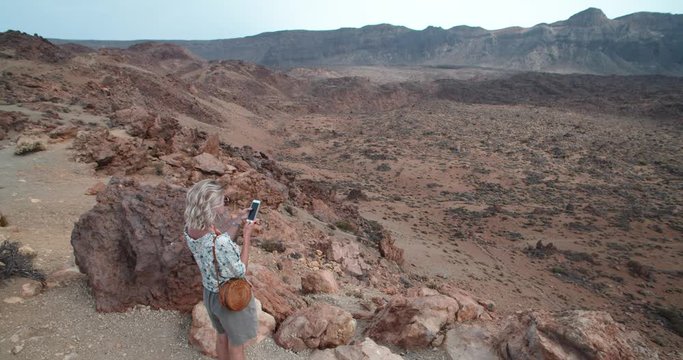 Modern blonde girl using smartphone standing on famous Teide volcano crater landscape. Tourist woman taking photos on smartphone admiring volcanic scenery. Teide National Park, Tenerife.