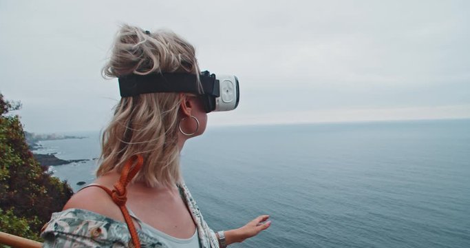 Young cheerful woman using VR-headset technology staying above blue ocean enjoying picturesque scenery of Tenerife. Virtual reality. People and technology.