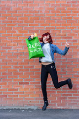 Smiling young girl in a jump with an eco bag with white text zero waste. The girl has red short...